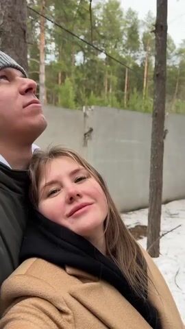 In a natural outdoor setting in Polazna, Russia, a man and a woman are seen together. The woman, with long brown hair and a slight smile, rests her head on the man's shoulder, looking directly at the camera. She is wearing a black hooded top and a light brown or camel-colored coat. The man, whose face is partially visible on the left, wears a dark jacket and a grey beanie, looking upwards.

The ground around them is covered with patches of snow, suggesting a cold season like winter or early spring. Behind them, a concrete wall is visible, separating the natural space from what appears to be a man-made structure or boundary. Tall pine trees characterize the environment, with their trunks forming a dense forest.

As the video progresses, the camera pans upwards, focusing on the canopies of the tall pine trees swaying, likely due to wind. The man's voice is heard, stating in Russian, "Ветер сильный" (The wind is strong), confirming the windy conditions affecting the trees. The sounds of nature, including wind, are audible. The overall mood is calm and natural, despite the mention of strong wind.