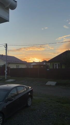 The media file captures a sunset scene in a residential area, likely in Bobrovsky, Russia. The sky is painted with warm hues of orange and yellow, with scattered clouds catching the last rays of sunlight. The setting appears to be a suburban neighborhood with houses featuring red-tiled roofs and dark brown fences. A black car is parked in the foreground, suggesting this is a view from a driveway or yard. Utility poles and power lines crisscross the sky, a common sight in such areas. The overall mood is peaceful and serene, characteristic of a calm evening at the end of the day. The time of day is clearly sunset, indicated by the low position of the sun and the warm, diffused light. There are no people visible in the frame, and the focus is on the natural beauty of the sunset and the quiet suburban landscape. No discernible text is visible in the media file.