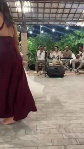 At what appears to be an outdoor wedding reception, a bride in a white wedding gown is dancing with her bridesmaids, who are wearing floor-length gowns in various pastel colors. The dance floor is made of light-colored patterned tiles. Behind the dancers, a live band of five musicians, all men, are playing instruments and singing into microphones. The band is set up with speakers and other equipment. Overhead, string lights illuminate the outdoor space, which is covered by a wooden roof structure. Some greenery, including trees and shrubs, can be seen in the background.