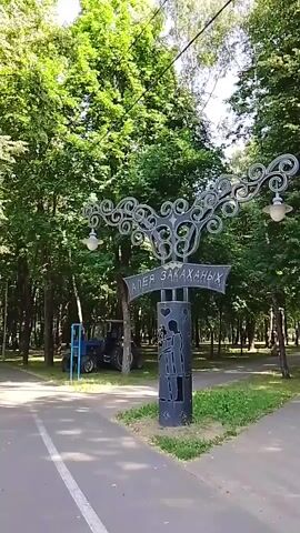 A wide shot shows a park on a sunny day. The camera is positioned at the edge of a paved path, looking towards a large ornamental lamppost. The lamppost is dark metal with intricate scrollwork at the top and two glass light fixtures. A rectangular sign is attached to the lamppost with Russian text. Below the sign, the lamppost is decorated with a silhouette of a couple embracing. To the left of the lamppost, a blue tractor is parked on a grassy area. Behind the tractor and lamppost, a dense line of tall, green trees forms a backdrop. Sunlight filters through the leaves, creating dappled light and shadows.