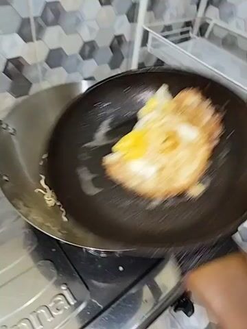 A person's hands are visible, holding a black frying pan with a fried egg in it, and a wooden-handled spatula. The frying pan is being tilted over a large metal wok that is on a gas stove. Inside the wok are instant noodles and some green vegetables. The person is adding the fried egg from the frying pan into the wok with the noodles and vegetables. The background shows a tiled backsplash and a white shelf with some items on it, including what appear to be onions and garlic.