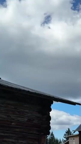 The video displays an exterior shot of traditional wooden buildings under a cloudy sky. The camera pans across the scene, revealing rustic log structures with dark roofs. In the foreground, a portion of a dark wooden building juts into the frame, its rough-hewn logs visible. As the camera moves right, more of the buildings come into view, including a small structure with windows and a more elaborate wooden church-like building with a domed roof topped by a cross. In the background, evergreen trees are visible against the bright blue sky, punctuated by large, grey and white clouds. The overall impression is one of an old, possibly historical, rural or religious settlement.