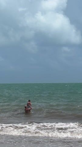 Two individuals stand in the ocean near the shore. One person stands deeper in the water, with the other person sitting on their shoulders. Waves gently break on the beach in the foreground. The ocean extends to the horizon, meeting a cloudy sky. The water appears green, and the sky is a mix of blue and gray.