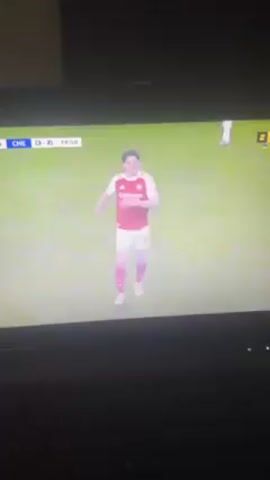 A laptop screen shows a soccer game between Chelsea and Arsenal. The score is Chelsea 3, Arsenal 2, and the time is 74:55. The camera then focuses on a man in a dark jacket standing on the sideline. The background is filled with other people in various clothing. The laptop's keyboard is visible in the foreground.