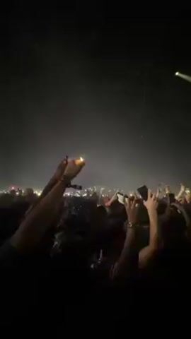 A large crowd of people is gathered in a dark indoor venue, likely a concert hall or arena. The scene is illuminated by stage lights and large screens, creating an energetic atmosphere. Above the crowd, triangular light fixtures are visible. The crowd is highly engaged, with many people raising their hands in the air, cheering, and recording the event with their phones.

A massive LED screen at the front displays vibrant animated art of a person with long, dark hair, wearing a red top and jewelry. The animation is dynamic, shifting colors and perspectives. The person in the animation has their hands outstretched, contributing to the grand scale of the visual presentation. The music and cheering indicate a live performance or DJ set. The overall mood is one of excitement and celebration.