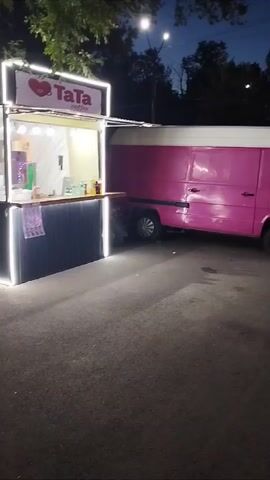 The video, likely filmed in Kara-Balta, Kyrgyzstan, captures a lively evening scene, possibly at a fairground or public gathering. The time of day is dusk or early evening, as indicated by the darkening sky and the illuminated objects.

The primary subjects are a vibrant pink food truck, branded "TaTa Coffee," and a row of brightly lit, child-sized amusement rides resembling small cars or vehicles. These rides are adorned with colorful lights, creating a playful and inviting atmosphere. Several people are visible, including a woman in a red shirt and denim shorts walking towards the camera, and other individuals interacting with the rides or walking in the background. A person pushing a stroller is also present, suggesting a family-friendly environment.

The setting appears to be an outdoor, paved area, possibly a street or a designated event space. Trees and overhead lighting contribute to the ambiance. The overall mood seems cheerful and energetic, with the colorful lights and the presence of people enjoying themselves. The juxtaposition of the "TaTa Coffee" truck, suggesting a place for refreshments, with the amusement rides implies an event where food and entertainment are offered.