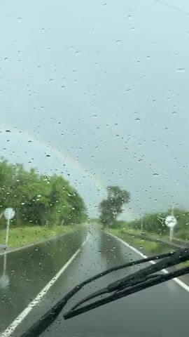 The video shows a view from inside a vehicle driving on a wet road. Raindrops are visible on the windshield. There is a rainbow in the distance. The road is straight and lined with trees on either side. The sky is overcast and gray. The road surface is dark and reflective due to the rain. There are white lane markings on the road.