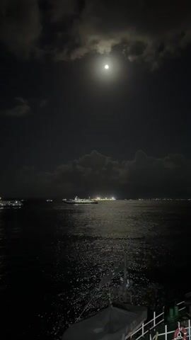 A nighttime video captures a full moon shining brightly in a cloudy sky over a dark ocean. The moon's reflection shimmers on the water's surface, creating a path of light. In the distance, several illuminated ships are visible on the horizon. In the foreground, the railing and part of a boat are visible, suggesting the video is filmed from another vessel. The overall atmosphere is calm and serene.