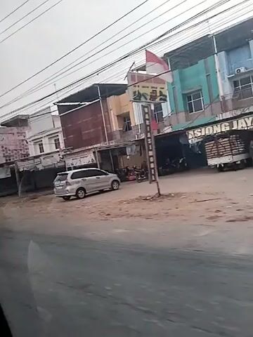 This is a video taken from a moving vehicle, likely a car, showing a street scene. On the right side of the street, there are several buildings that appear to be shops or businesses. One building has signs that say "KARTU" and "MERDEKA MOTOR SERVICE & SPAREPART". There are several motorcycles parked in front of one of the shops. A silver van is parked on the street. A black SUV is in the foreground, moving from left to right across the screen. Further down the street, a green bus is passing by, moving in the opposite direction. The street is paved, and there are overhead power lines strung across the scene. The sky appears overcast.