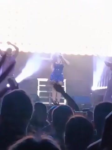A woman is performing on stage in front of a crowd. She is wearing a short blue dress, has blonde hair, and is using a microphone. Behind her is a backdrop of lights. The crowd is raising their hands and cheering. Confetti is falling. The stage appears to be made of metal and has multiple tiers.