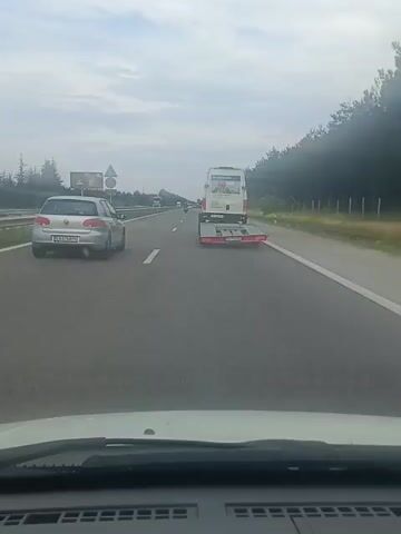 This is a first-person perspective video taken from a car driving on a highway, likely in Tserovo, Bulgaria. The car is in the right lane, following a white van with a trailer and a silver car in front of it. The van has some text on its side that appears to be a company name and possibly a slogan, but it is partially obscured and difficult to read clearly. The highway is flanked by trees on the right and a grassy embankment with a fence on the left. There are other vehicles visible in the distance, including trucks and cars. The sky is overcast, suggesting a cloudy or possibly rainy day. The video appears to be taken during daylight hours. The overall mood is that of travel and a typical highway commute. There is no indication of any specific event or emotional state beyond that of a driver navigating a road.