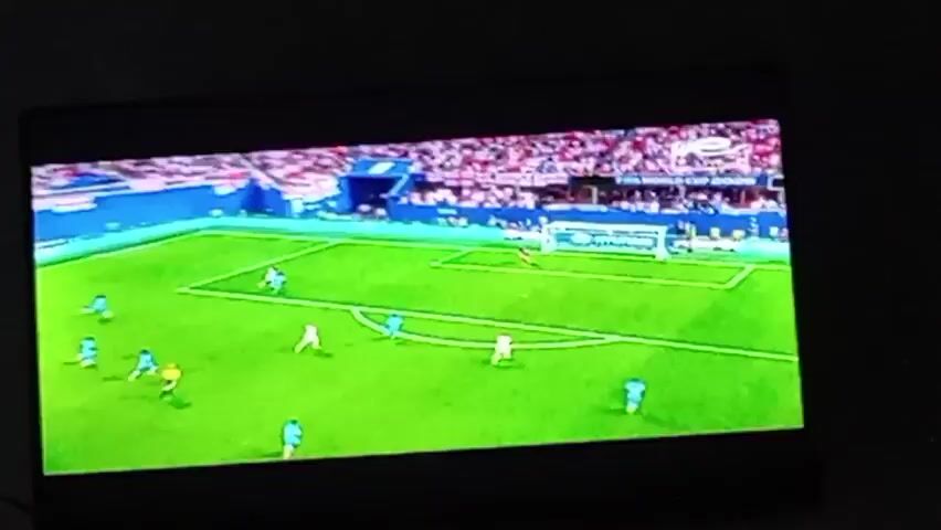 This is a video recording of a soccer match being played on a bright, sunny day, likely in the afternoon given the lighting. The scene is a vibrant green soccer field with clear white boundary lines. Spectators in a stadium, wearing red and white, fill the stands behind the field. The match features players in two distinct uniforms: one team in light blue jerseys and the other in white jerseys. The game appears to be in progress, with players actively engaged in play near the goal. The players in blue are spread out across the field, with one near the goal and others positioned further back. The white-jerseyed players are also scattered, with one near the goal and another in the middle of the field. The overall atmosphere seems to be one of intense competition. Visible text on the screen includes "FIFA WORLD CUP" and "LIVE".