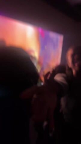 The video shows a group of people standing in front of a large screen displaying a colorful image of a young girl. The people are mostly obscured by the darkness, but some are holding cups and appear to be celebrating or enjoying themselves. The environment seems to be an indoor space, possibly a party venue or event hall. The overall atmosphere is lively and festive.