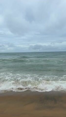 This video shows a tranquil beach scene with waves gently rolling onto the shore under a cloudy sky. The water is a greenish-blue color, and the sand is a light brown. The waves create a foamy white line as they meet the beach. There are no people or man-made objects visible in the frame. The overall atmosphere is peaceful and serene.