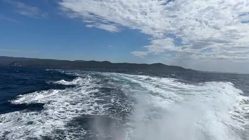 The video shows a view from the stern of a boat moving through the ocean. The water is choppy, with white foam and a long wake trailing behind the boat. In the distance, a green, hilly coastline with trees is visible under a bright blue sky with scattered white clouds. The sun appears to be high in the sky. There are no people or other objects clearly visible in the frame. The camera is stationary relative to the boat, capturing the motion of the water and the distant landscape.