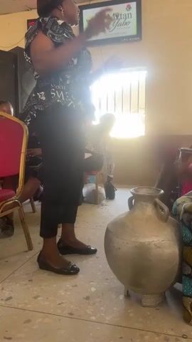 A woman stands in front of a large, clay pot, speaking into a microphone. She wears a black and white top, black pants, and black shoes. Behind her, people are seated on chairs. A monitor displays the words "Mtan Yabo." A woman to the right of the clay pot is playing an instrument. The room is well-lit, but bright sunlight streams in through a barred window.