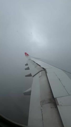 The video shows the wing of a plane, seen from a window seat, flying through heavy fog. The wing is white with some black markings, and has the Turkish Airlines logo on the tip. The fog is dense and obscures any view of the ground or sky beyond the wing.