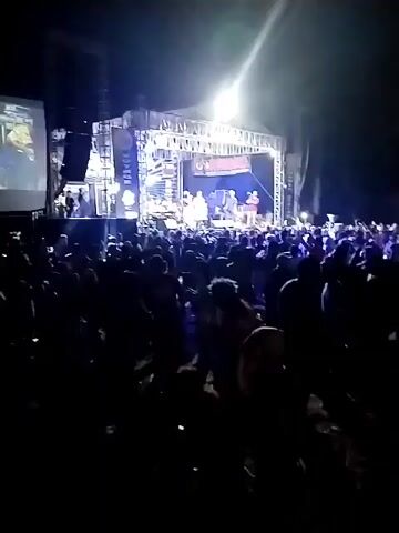 A live music concert is taking place outdoors at night. A band performs on a brightly lit stage, with a large crowd gathered in front of it. The audience is densely packed and appears to be enjoying the performance, with many people raising their hands in the air. A large screen is visible on the left side of the stage, displaying content from the performance. The overall atmosphere is energetic and festive, with spotlights illuminating the stage and the crowd.