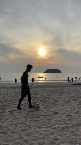 A video captures a sunset scene on a sandy beach in Tambon Karon, Thailand. In the foreground, a young man, silhouetted against the setting sun, is playing football. He is seen kicking the ball, then walking away from the camera. The sun is low on the horizon, casting a warm, golden glow across the sky and reflecting on the ocean's surface. In the background, a small island is visible in the distance. Numerous other people are scattered along the beach and in the water, enjoying the evening. Some boats are also seen in the water. The overall atmosphere is peaceful and leisurely, typical of a beach at dusk.