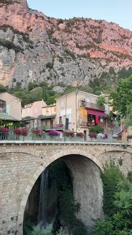 This video captures a serene scene in Moustiers-Sainte-Marie, France, during what appears to be the golden hour. The setting is a charming village nestled at the base of a majestic, rocky mountain that is bathed in warm, rosy light, suggesting either sunrise or sunset. A stone bridge, adorned with vibrant potted flowers in shades of pink, purple, and white, spans over a cascade of water, creating a picturesque waterfall that disappears into lush greenery below.

Several buildings with traditional European architecture are visible on the slope leading up from the bridge. Some of these structures feature balconies and outdoor seating areas, hinting at cafes or restaurants where people are enjoying the evening. Two individuals can be seen on one of the balconies, their figures silhouetted against the warm light. Further along the bridge, a few more people are strolling or pausing, adding a sense of gentle activity to the tranquil atmosphere. The weather appears pleasant, with clear skies overhead. The overall mood conveyed is one of peace and natural beauty, enhanced by the soft lighting and the vibrant colors of the flowers against the rugged mountain backdrop. No visible text is present in the video.