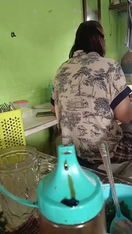 The video shows a view from behind a person wearing a patterned shirt. The person is seated at a table in a room with light green walls. In the foreground, there is a glass, a turquoise-lidded bottle, and a bowl with a spoon. In the background, a woman and a young girl are sitting at another table. The woman is looking down at something on the table, and the girl is facing towards the camera. The room appears to be a simple eatery or home setting. There is a mirror on the wall, a fan on the ceiling, and a calendar hanging on the wall. Sunlight is coming in from an open doorway in the background.