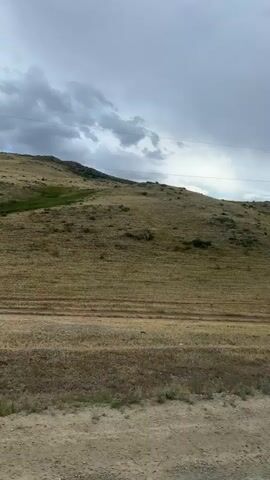 The video captures a view from a moving vehicle on a dirt road, looking out towards a rolling, grassy hillside under a cloudy sky. A single utility pole with power lines is visible on the left side of the frame. The terrain is predominantly covered in dry, golden grass, with some patches of greener vegetation on the hillside, possibly indicating cultivated fields or different types of plants. A winding dirt path or road is visible on the hillside. The sky is filled with dramatic, dark clouds, suggesting approaching weather, with some lighter patches and sunlight breaking through in the distance. The overall impression is of a rural, possibly remote landscape.