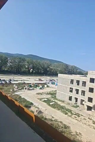 The video shows a construction site with a building under construction in the foreground. The building appears to be made of concrete and has several windows. In the background, there are residential buildings, a playground, and a wooded hill. The sky is clear and blue.