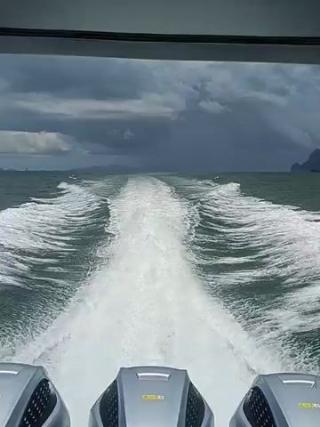 This media file is a first-person perspective video captured from the back of a speeding boat. The boat appears to have multiple outboard motors, visible at the bottom of the frame. The boat is leaving a prominent white wake on the choppy, dark green water. In the distance, the horizon shows a hazy, overcast sky with dark, ominous clouds, suggesting either an approaching storm or a recent downpour. The sea extends to the horizon where a faint silhouette of land, possibly islands or a coastline with distinctive rock formations, can be seen. The overall atmosphere is one of movement and power, with the churning water and the distant, brooding sky contributing to a sense of journey or adventure. While no people are directly visible, the perspective implies someone is operating or traveling on the boat. The location cues, like the rocky islands in the distance, are consistent with the coastline of Ko Panyi, Thailand, a region known for its dramatic limestone karsts. The time of day is unclear due to the overcast conditions, but the lighting suggests it is daytime.