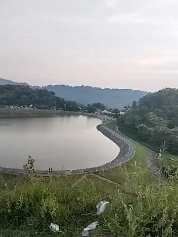 The video presents a panoramic outdoor view of a large reservoir in Jambeyan, Indonesia, likely during late afternoon or early evening under a hazy sky. In the immediate foreground, green grass is visible, scattered with some litter. A man wearing a yellow short-sleeved shirt and dark pants enters the frame from the left, walks briefly, then turns to face the reservoir and spreads his arms wide, gesturing towards the expansive scenery. The reservoir, with its calm, reflective water, curves prominently in the midground, bordered by a paved road or dam wall. Small figures of people can be seen along this curved structure in the distance. Beyond the reservoir, a series of verdant, tree-covered hills rise, completing the scenic landscape.