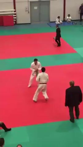 Two competitors are engaged in a judo match on a red and green mat. One competitor wears a white judogi with a red belt, while the other wears a white judogi with a green belt. They are grappling, attempting to gain an advantage. A man in a suit stands near the edge of the mat, observing the match. In the background, several people are seated on a blue mat, also watching. A chair is placed near the edge of the mat. The setting appears to be an indoor sports facility, possibly a gymnasium.