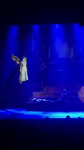 The image captures a theatrical performance bathed in dramatic blue stage lighting. The scene appears to be set on a stage with tiered platforms and a dark, possibly ancient or gothic, backdrop.

The central figure is a woman dressed in a flowing white gown and adorned with large, golden angel wings. She is suspended in the air, suggesting a special effect or harness, and has long blonde hair. Her expression is serene, and she seems to be observing the scene below.

On the platforms below, two figures are visible. One is lying down, seemingly relaxed or perhaps in a state of repose. The other figure is seated, facing away from the viewer, adding to the mysterious atmosphere.

The overall mood is ethereal and dramatic, enhanced by the strong blue lighting that casts long shadows and highlights the figures. The presence of a chandelier-like structure in the background further suggests a theatrical setting. The audience is dimly visible in the foreground, indicating this is a live performance.

There is no visible text in the image. The specific event or narrative being depicted is not immediately clear from the visual alone, but the angelic figure and the subdued, almost somber poses of the other characters hint at a story with symbolic or mythical elements. The time of day and weather are irrelevant as it is an indoor stage production. The location context of Шебунино, Russia, implies the performance is taking place there, but provides no visual cues within the image itself.