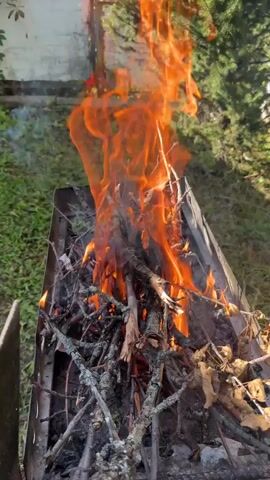 A close-up video shows a campfire burning in a metal grill outdoors. The flames are bright orange and yellow, with some smoke visible. The grill is filled with dry branches and leaves. The background is blurred, but it appears to be a grassy area with some trees and a white structure. The camera is angled downwards towards the fire.