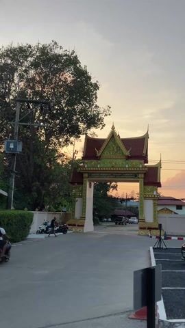 The video depicts a street scene featuring a traditional Thai archway, likely marking an entrance to a temple or significant area. The archway is elaborately decorated with gold and green accents, a red roof, and Thai script. The sky behind the arch is a gradient of light pinks and oranges, indicating either sunrise or sunset. A couple of people are riding a motorcycle across the frame from left to right, and a pedestrian is walking on the left-hand side of the road. Other parked motorcycles are visible on the left, and several cars are parked or passing through the archway. On the right, a gray car is parked in a designated parking spot. The overall atmosphere is calm and serene, reflecting the late evening light.