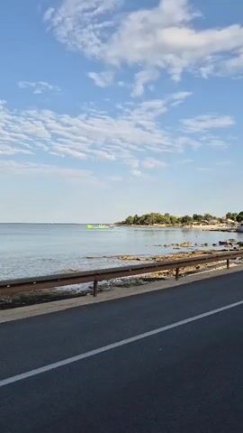 A video shows a body of water, likely the sea, under a bright blue sky with scattered clouds. In the foreground, a paved road with a white line is visible, separated from the water by a brown metal guardrail. The water appears calm and reflects the sky. Along the edge of the water, there is a rocky shoreline. Further in the distance, a small green structure or inflatable object can be seen in the water, and a tree-lined coast is visible. The overall scene suggests a coastal road with a view of the sea.