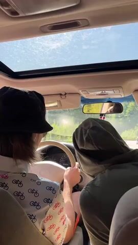 The video shows the interior of a car from the perspective of a passenger in the back seat. The car has a sunroof that is open, and the sun is shining through it. Two people are visible in the front seats. The person in the driver's seat is wearing a black bucket hat and a white shirt with a pattern of bicycles on it. They are holding the steering wheel, and their hands are visible. The person in the passenger seat is wearing a green hooded sweatshirt and their back is to the camera. They appear to be looking out the window. The car is driving on a road surrounded by trees, and the sun is glinting off the windshield. A rear-view mirror is visible, and a beaded string with a small phone attached hangs from it. The interior of the car appears to be beige.