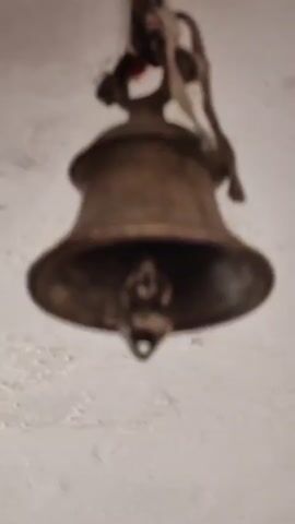 A bronze bell hangs from a rope attached to a white ceiling. The bell is old and tarnished, with some debris inside. A small, blurry object is seen in the periphery of the video.