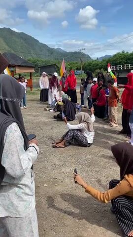 A group of women and children are gathered outdoors for what appears to be a game. In the foreground, two women, one in a light-colored hijab and patterned pants, and another in an orange top and striped pants, are holding phones, seemingly recording or observing. The woman in the foreground left is partially obscured. In the middle ground, several women are sitting on the ground in a line. One woman, wearing a black hijab and blue top, is reaching for a knife on the ground. Another woman, in a light green hijab and patterned pants, is in a similar position. A third woman, in a white shirt and patterned pants, is also seated. Behind them, a group of people, including women in hijabs and children, are standing and watching the game. Flags are visible in the background. The environment is outdoors with a dirt ground, trees, and mountains in the distance under a partly cloudy sky.