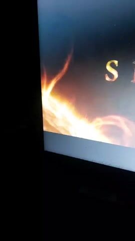 The video shows a close-up of a screen displaying the title "SKY DANCE". The title is rendered in a metallic, golden font with a fiery effect. Behind the text, there is a cosmic background with swirling nebulas and explosions of light, suggesting a celestial or fantasy theme. The lower part of the screen has a white border, indicating it might be a computer monitor or a television. The surrounding environment is dark, suggesting the video is being watched in a dimly lit room. There are no people visible in the frame.