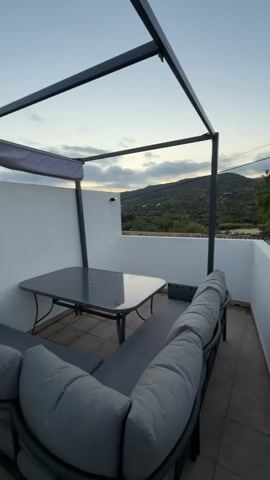 The video shows an outdoor patio area with a gray outdoor sofa and a gray table with a glass top. A metal pergola covers part of the patio. In the background, there are rolling hills covered with green vegetation and some buildings with terracotta roofs. The sky is cloudy and appears to be either dusk or dawn. There are also several small spotlights on the ground.