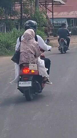 A man wearing a black helmet and a white shirt rides a red scooter. A woman wearing a light-colored hijab and a patterned outfit sits behind him. She is carrying two bags, one black and one beige. The scooter has a license plate that reads "BL 4935ZBT". Another scooter and rider are visible in the background. The scene appears to be outdoors on a paved road, with vegetation and buildings in the background.