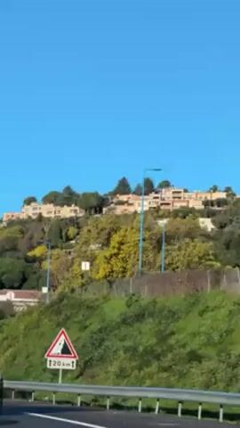 The camera, moving from a vehicle, captures a roadside view on a sunny day. The scene features a green, overgrown hillside dotted with trees, punctuated by streetlights. Several buildings, appearing residential, are visible at the hilltop. Road signs are prominently displayed: one is a triangular warning sign showing a falling rocks symbol, marked with "20 km," and another is a rectangular sign indicating "FAYENCE," "LES ADRETS DE L'ESTEREL," and "D 39" with a "10" above it. The sky is a clear, solid blue.
