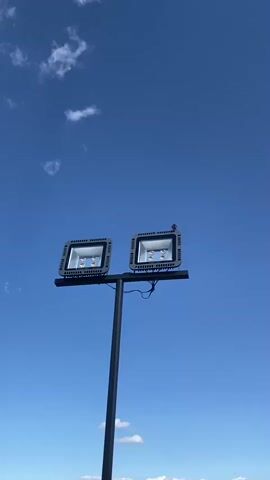 A small bird is perched on the edge of a bright, square LED floodlight. The floodlight is mounted on a metal pole along with a second, identical floodlight to its left. Both floodlights are silver with a textured border and have four glowing orange LED bulbs visible inside. The bird, which appears to be a sparrow or similar small passerine, is facing to the right and is a dappled brown and white. It is positioned on the upper right corner of the rightmost floodlight. The floodlights are attached to a horizontal metal bar, which is then attached to a vertical gray pole. Wires are visible trailing down from the floodlights.

The scene is set against a clear, vibrant blue sky with a few wispy white clouds scattered towards the top of the frame. The sun is bright, casting a strong light on the floodlights and the bird. The time of day appears to be daytime, likely late morning or early afternoon given the brightness and the clear sky. The setting appears to be outdoors, possibly a public space, parking lot, or sports field where such floodlights would be installed. The image evokes a sense of peacefulness and nature interacting with man-made structures. There is no discernible text in the visible portion of the image beyond the faint "EUROLIGHT" label on the floodlight. The location context is Artashavan, Armenia.