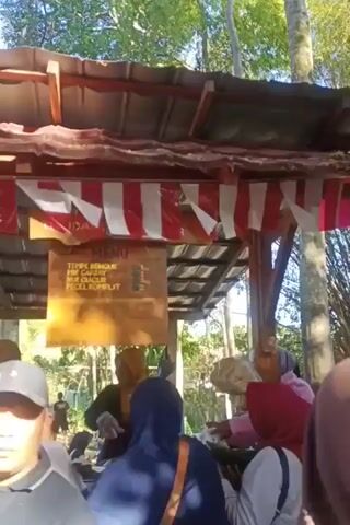 A group of people are gathered around a food stall. The stall has a wooden roof with red and white striped fabric draped along the edge. A wooden menu board is hanging from the roof. The menu lists "TEMPE BENGUK", "MIE GORENG", "AYAM GORENG", "NUS GULUR", and "PECEL KOMPLIT". Several people are wearing hijabs. One person in the foreground is wearing a black baseball cap and a black polo shirt. Another person in the foreground is wearing a grey hijab and a blue jacket. The environment appears to be outdoors, with trees visible in the background. The lighting suggests it is daytime. Some people appear to be ordering food or waiting for their orders.