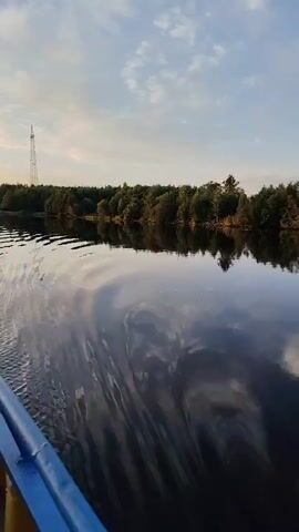 A calm lake reflects a treelined shore and a blue sky with scattered clouds. A white buoy is visible on the right side of the frame. The video appears to be shot from a moving watercraft.