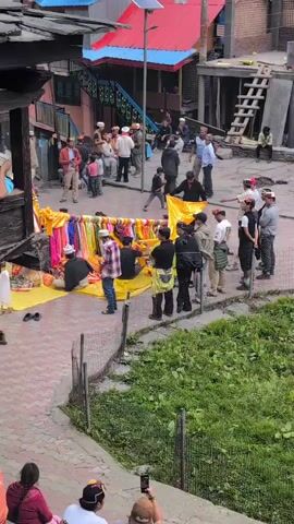 A high-angle shot shows a village square with several people gathered. In the center of the square, a group of people are arranging colorful cloths over a structure. Some individuals in the crowd are observing or participating in the arrangement. In the foreground, a few people are seated on steps, with one person filming the event with a smartphone. The background reveals multi-story buildings with vibrant colored roofs and a backdrop of lush green mountains. The overall scene suggests a local event or gathering taking place in a mountainous region.