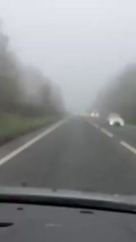 The video shows a view from inside a car driving on a road with heavy fog.  The road has white lines marking the lanes, and there's some greenery visible on the sides. Other cars are visible in the distance, their headlights appearing as blurred blobs. Water droplets are visible on the windshield.  The dashboard of the car is in the foreground.
