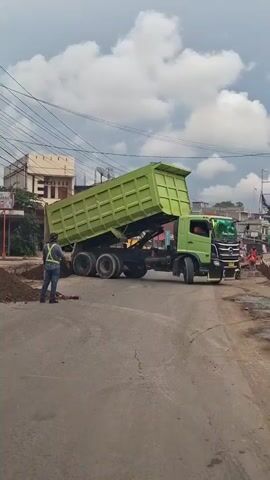 A dump truck with a green bed is shown from the side, parked on a dirt road with a person standing near the rear wheel. The truck's bed is raised, appearing to be in the process of dumping its load. Another person is visible in the driver's seat of the truck. The environment appears to be an outdoor construction or roadside area with buildings and power lines in the background. The sky is cloudy.