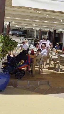 The video shows an outdoor seating area of a restaurant or cafe. People are seated at tables under a white canopy. In the foreground, a stroller is parked next to a table where a man and a boy are seated. The man is wearing a white t-shirt and sunglasses, and the boy is wearing a light-colored outfit. Behind them, other patrons are visible, some eating and others conversing. The environment appears to be sunny with some shadows cast by the canopy and the surrounding structures. The overall atmosphere suggests a casual dining experience.