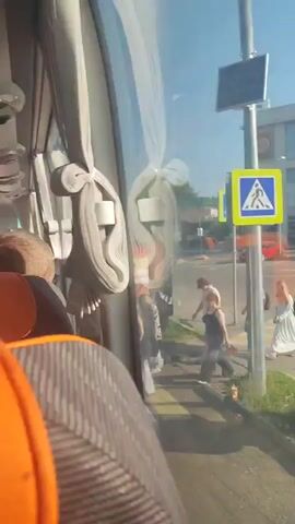 The video shows the interior of a bus. Orange seats are visible in the foreground, with gray and black patterned seats behind them. The ceiling of the bus has air vents and overhead storage compartments. Curtains are drawn along the windows, and sunlight streams through. Outside the window, people can be seen standing on a sidewalk next to a road, and a yellow road sign with a blue border is visible. The bus appears to be either stopped or moving slowly.