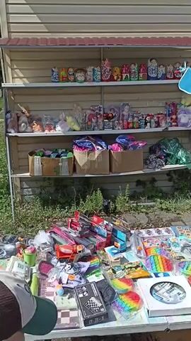 The video shows an outdoor market stall selling various items, primarily toys and bath accessories. The stall has multiple shelves displaying matryoshka dolls, stuffed animals, toy boxes, and other toys. In front of the stall, various items are laid out on a table, including toy cars, board games, and fidget toys. Hanging from the stall's awning are colorful bath sponges or loofahs. Two young girls are browsing the items at the stall. One girl, facing away from the camera, wears a baseball cap and a t-shirt. The other girl, also facing away, is wearing a white t-shirt with black stripes. The environment appears to be a sunny day, with some greenery visible in the background.